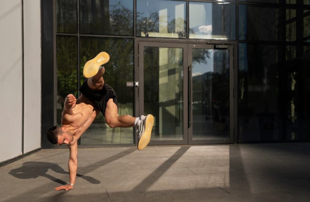 A man performs a one-handed handstand in a sunlit urban setting