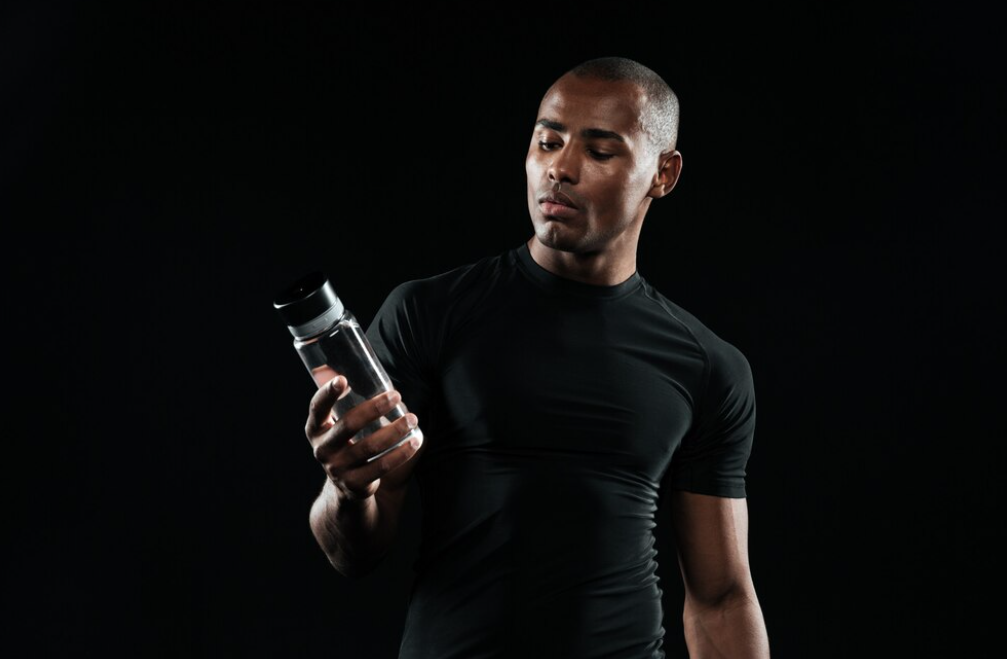 man in black t-shirt holds a bottle of water and looks at it on a black background