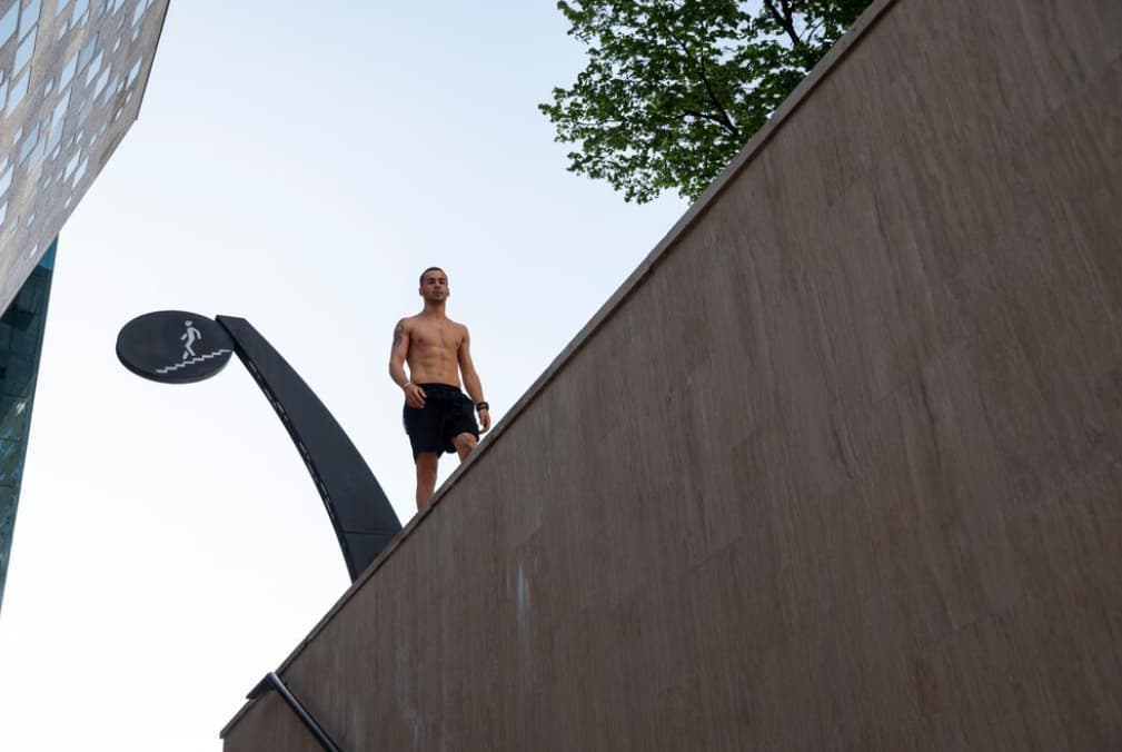 Shirtless athlete stands atop a high wall, looking down
