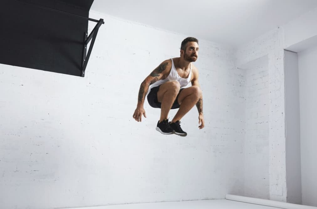 Tattooed man in a white tank top jumping indoors, with a white wall backdrop